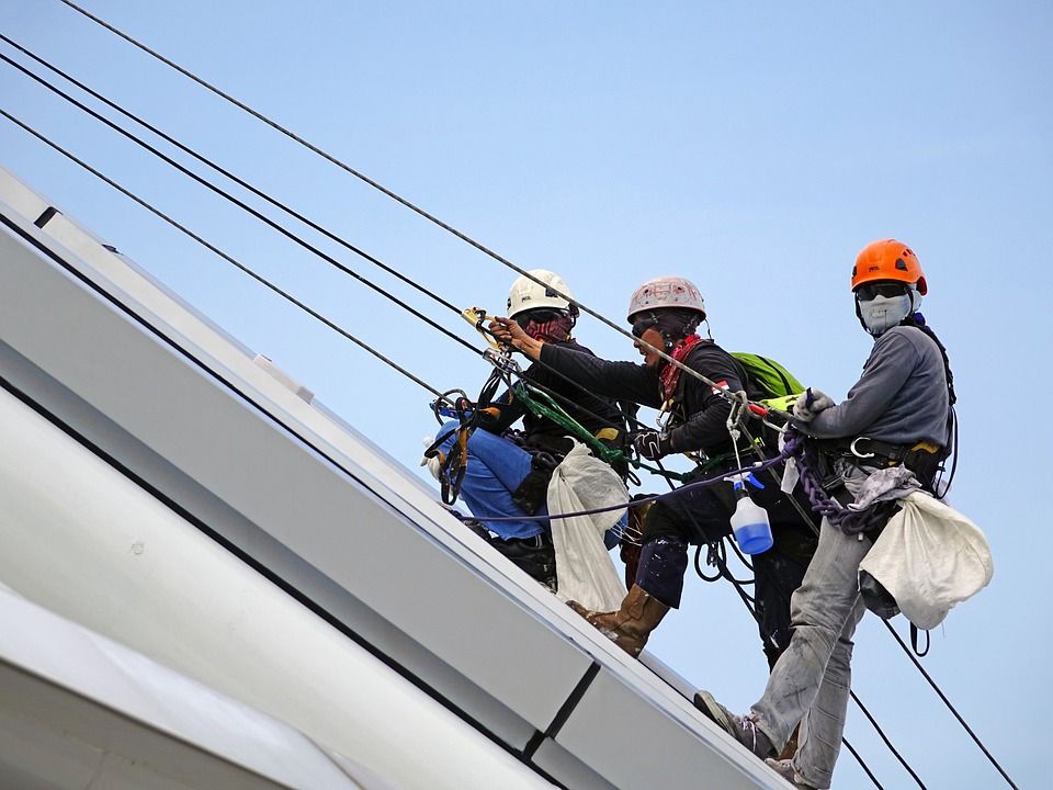 Trois ouvriers harnachés et suspendus à une corde travaillent sur un toit.